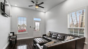 A spacious living room with a dark leather sofa, a matching ottoman, and a ceiling fan. There are two windows with blinds, one showing a red car outside.