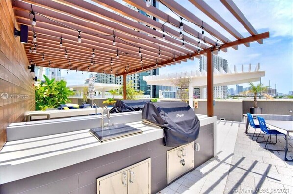 Un área de cocina al aire libre con una parrilla, taburetes de bar y una pérgola con luces colgantes.