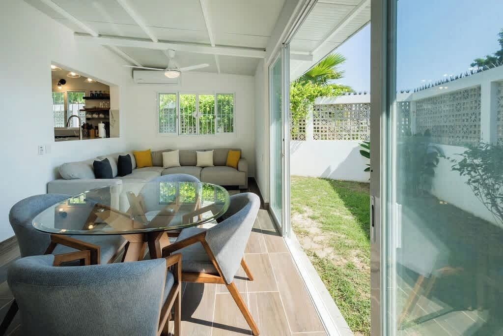 A modern living room with a glass table and chairs, a sofa with cushions, and a view of a garden through large sliding glass doors.