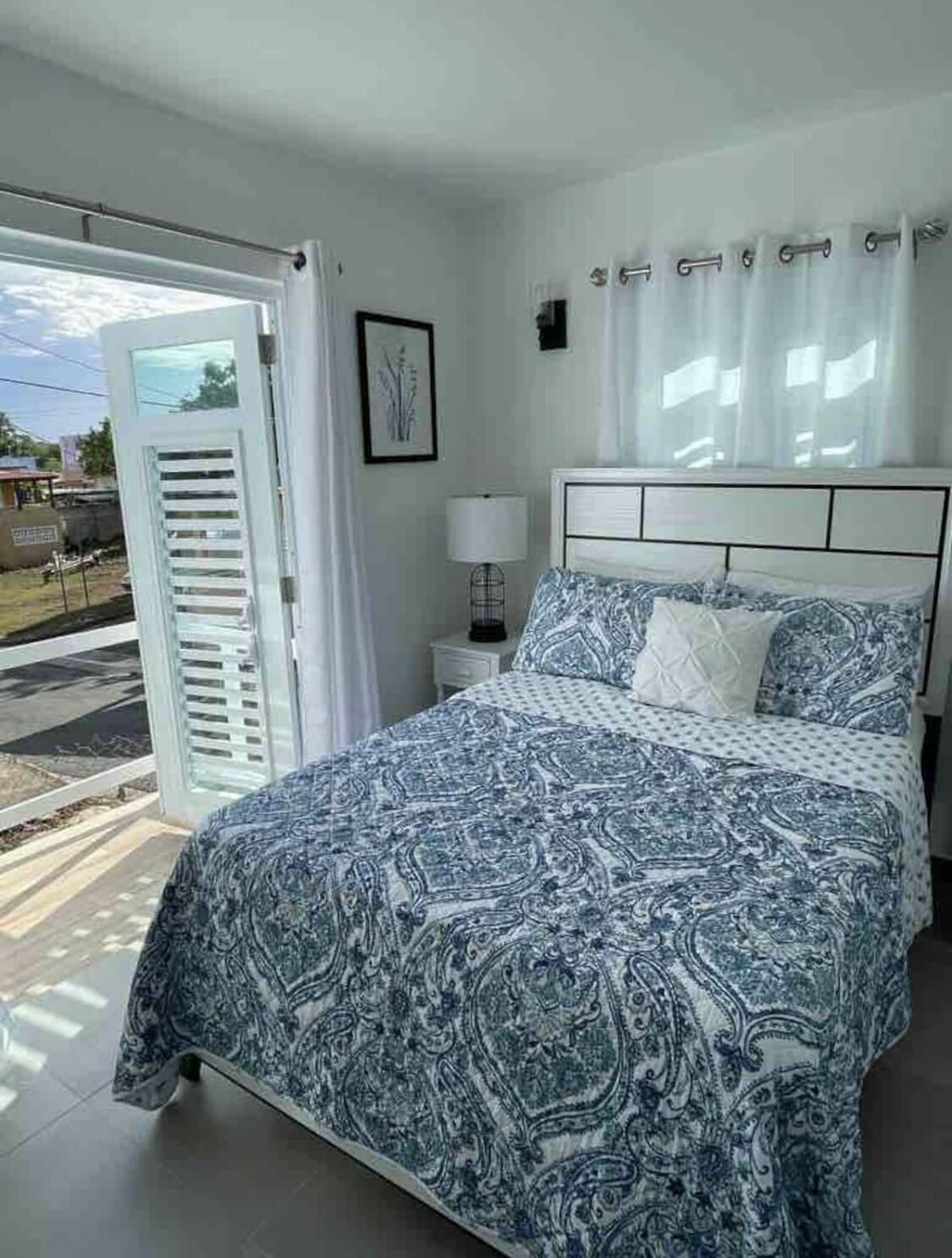 Une chambre à coucher comprenant un lit, une table de chevet, une lampe, une fenêtre avec des rideaux et une porte donnant sur l’extérieur.