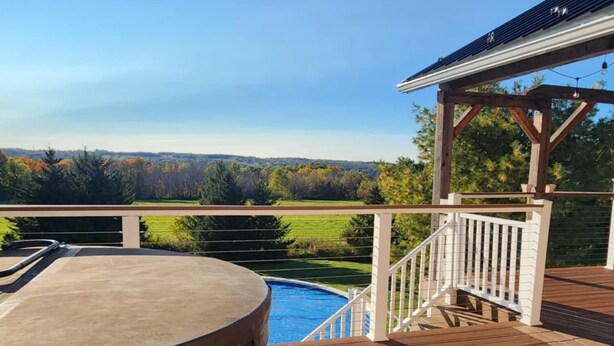 A wooden deck with a hot tub, a pool, and a gazebo overlooking a green landscape.
