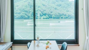 Ein Zimmer mit großem Fenster mit Blick auf einen See und Boote, ein Tisch mit Blumen und eine Lampe auf der Fensterbank.