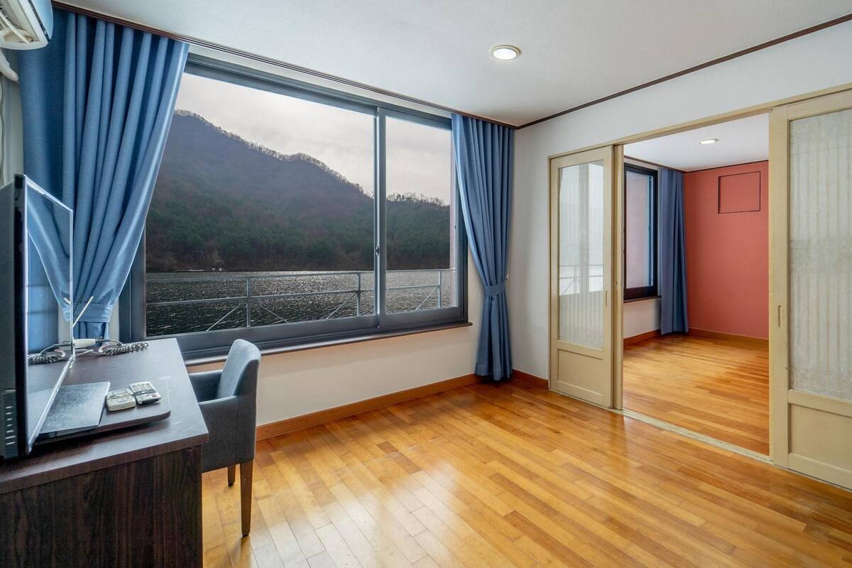 Una habitación con piso de madera, un escritorio con una silla, un ventanal con cortinas azules y vistas a una montaña y al agua.