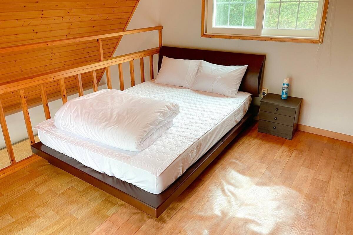 Un dormitorio con cabecera de cama de madera, ropa de cama blanca, una mesita de noche con una botella y una ventana con vistas a zonas verdes.