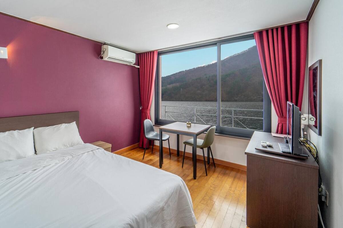 Habitación de hotel con cama, escritorio con silla, una mesita y un ventanal con vistas al agua y colinas.