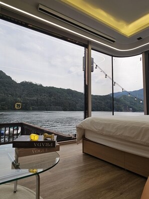 A modern bedroom with a large bed, a glass table with a book, and a view of a lake and forest. - Gapyeong Leupuang Pension (Gapyeong)