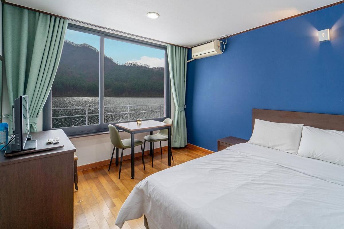 Habitación de hotel con cama, escritorio y silla. Hay una ventana con cortinas, unidad de aire acondicionado y vista a un cuerpo de agua y colinas.