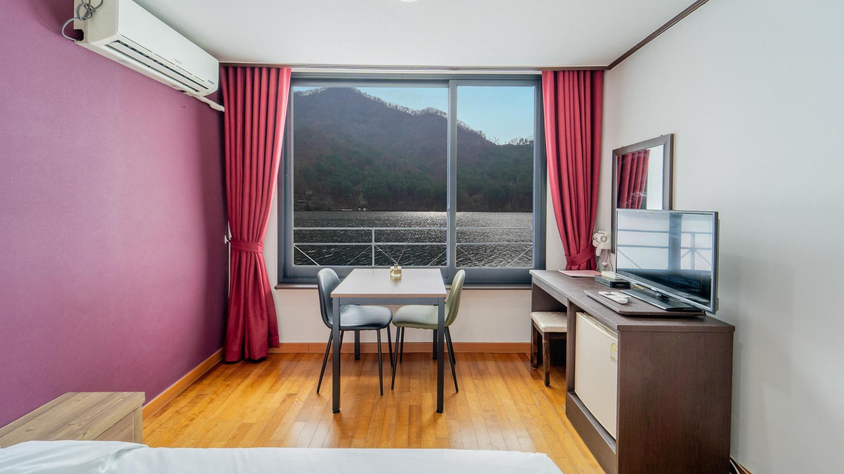A hotel room with a large window, a desk with a TV, a chair, and a bed.