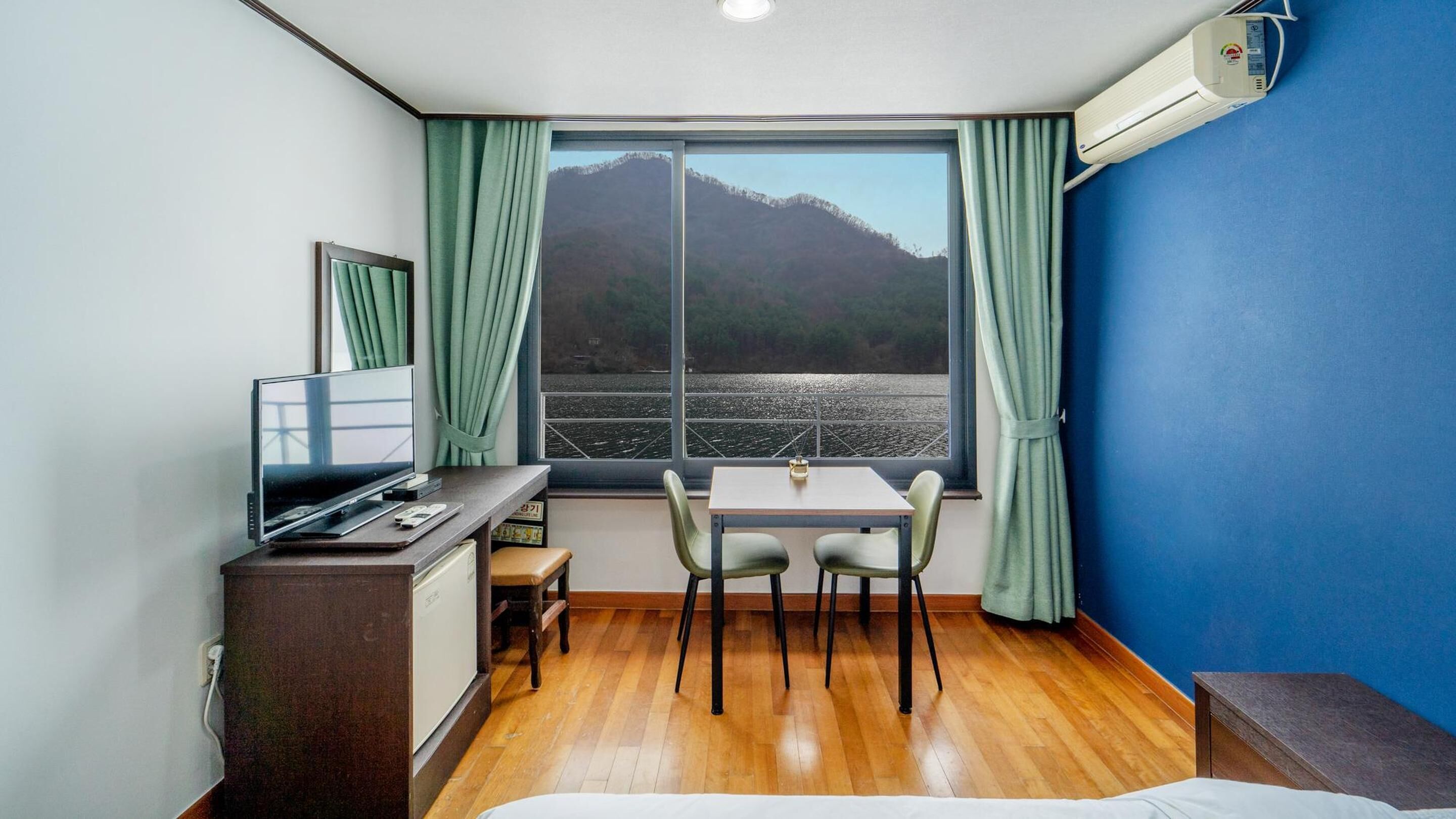 Ein Zimmer mit Bett, Schreibtisch mit Computer, Stuhl, Tisch und Fenster mit Blick auf Berge und Wasser.