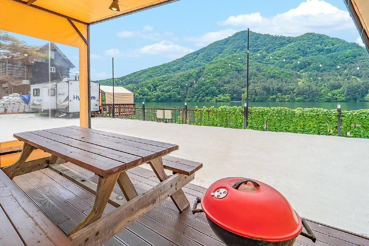 Una mesa de picnic de madera y una parrilla roja en una terraza con vista a un lago y una montaña.