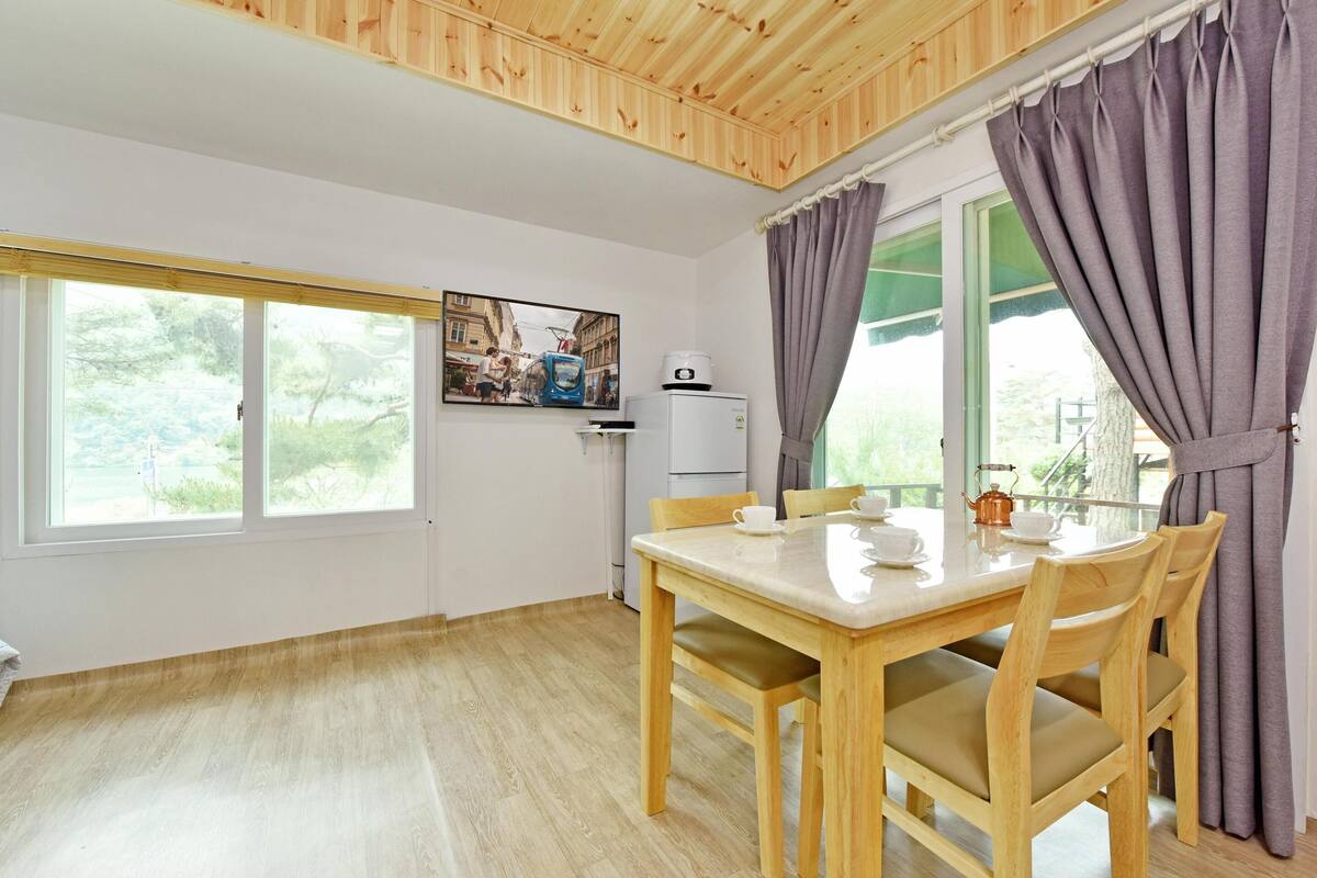 Una habitación con techo de madera, una mesa de comedor con sillas, una ventana con cortinas y una pequeña kitchenette.