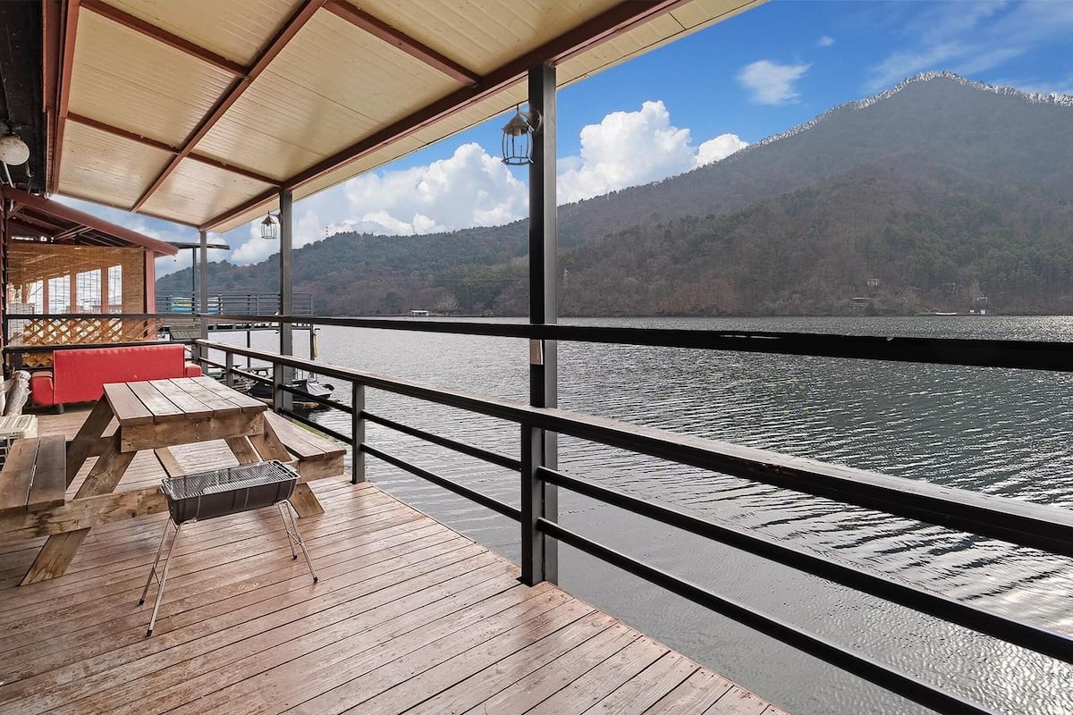 Una terraza de madera con mesas de picnic y un banco rojo, con vistas a un lago y montañas al fondo.