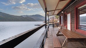 Eine Holzterrasse mit Geländer, die auf einen stillen See blickt, mit überdachtem Sitzbereich und Blick auf entfernte Berge.