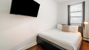 Een slaapkamer met een bed, een wandtelevisie, een bedlampje en een raam met jaloezieën.