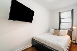 Un dormitorio con cama, televisor montado, lámpara de noche y ventana con persianas.