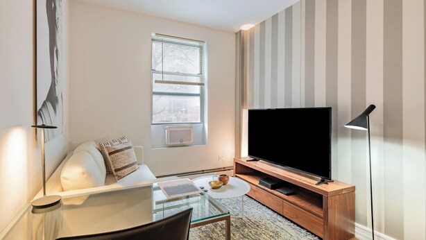 Een moderne woonkamer met een glazen tafel, een witte bank, een flatscreen televisie op een houten standaard, en een gestreepte wand.