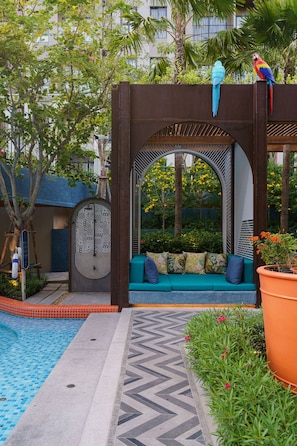 A pool area with a blue-tiled floor, a wooden pergola with a blue sofa, and a large orange planter.