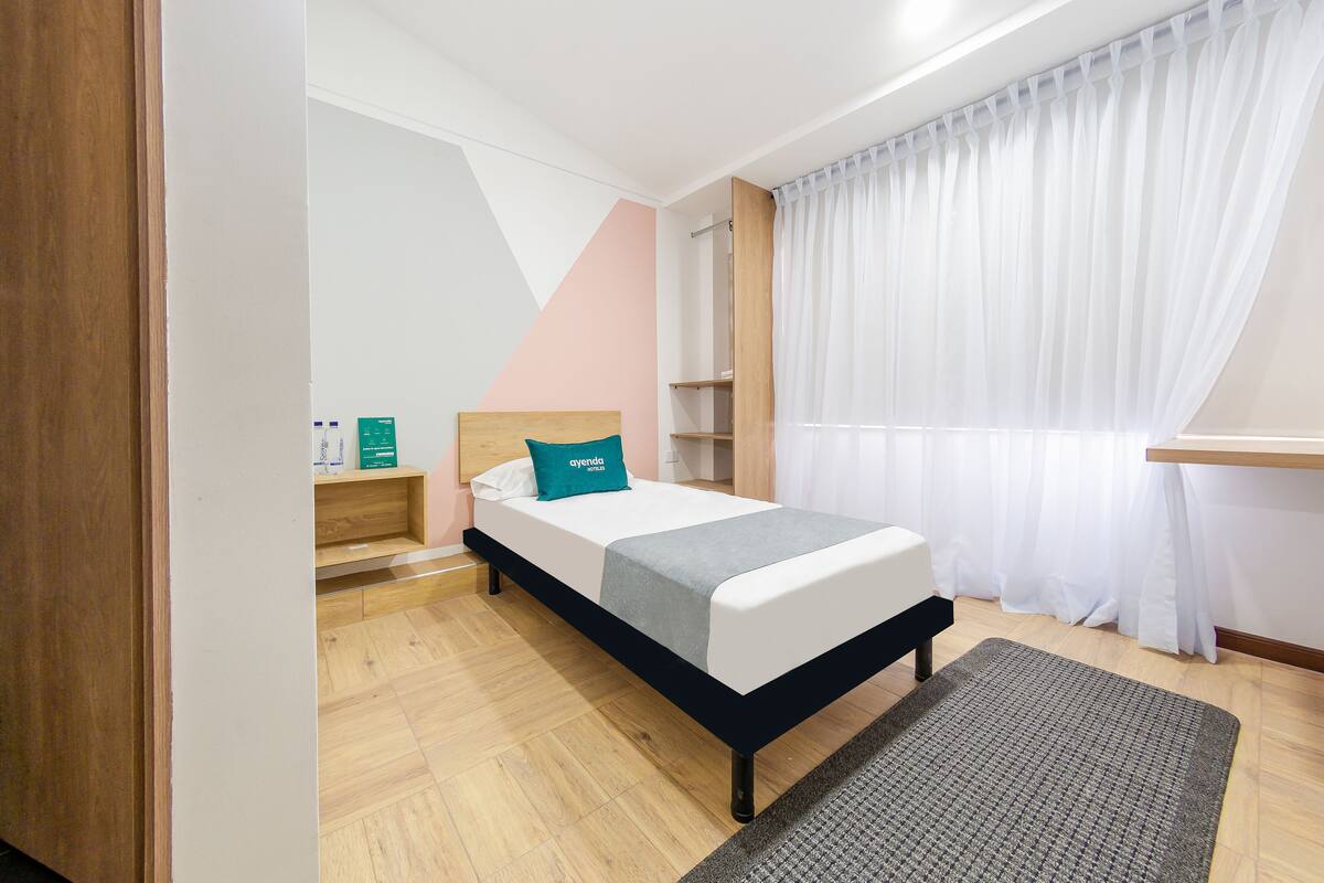 A bedroom with a bed, a bedside table, a nightstand, a window with curtains, and a wooden floor.