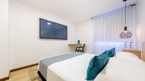 A neatly arranged hotel room with a bed, a desk, a chair, a TV mounted on the wall, and a window with curtains.