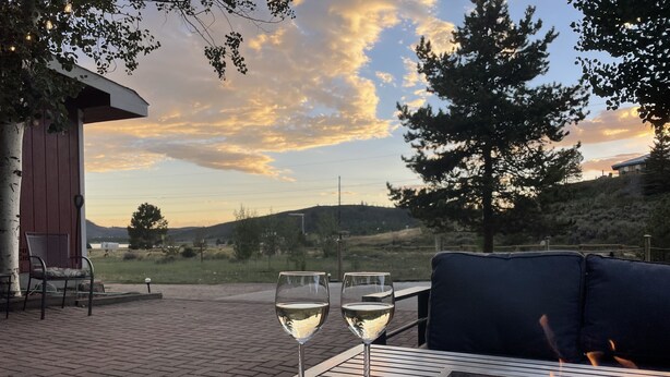 A patio with a fire pit, two glasses of white wine, and a view of the sunset over a hilly landscape.