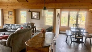 A cozy cabin interior with a wooden ceiling, a sofa, a dining table with chairs, and a kitchen area with a bar stool.