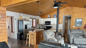 A modern kitchen with a refrigerator, microwave, and stove. There is a bar area with stools and a wooden counter. The living room has a gray sofa and a fireplace.