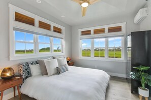 A bedroom with a large bed, two windows with views of a green landscape, a ceiling fan, and a potted plant.