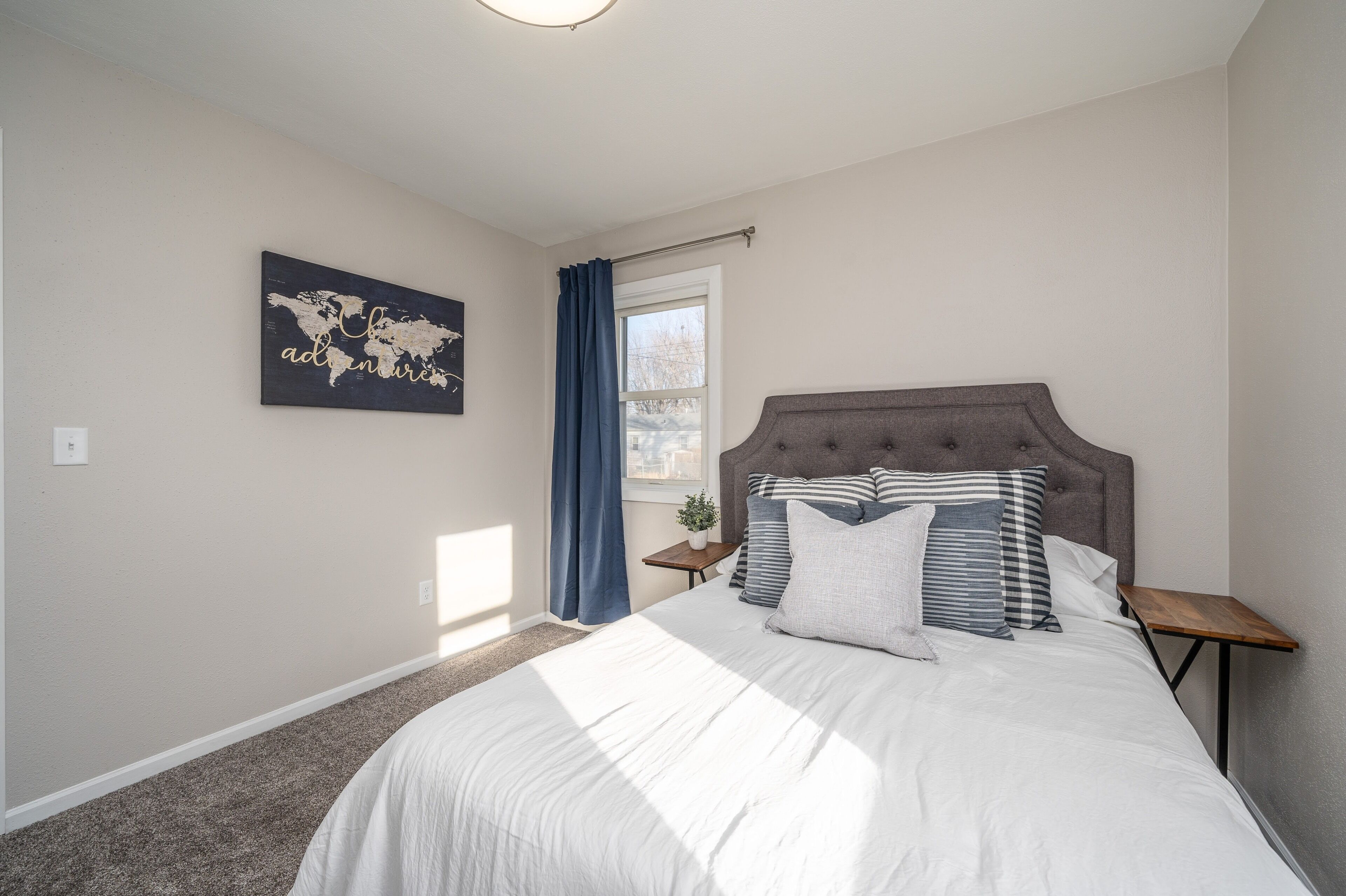 Une chambre avec un grand lit, une tête de lit, une table de chevet, une fenêtre avec des rideaux et une œuvre d’art représentant une carte du monde accrochée au mur.
