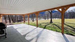 A covered patio with a concrete floor and wooden beams, overlooking a grassy area with trees and a mountain in the background. - The River House: stone section built in 1790, borders the scenic Juniata  River! (Mifflintown)