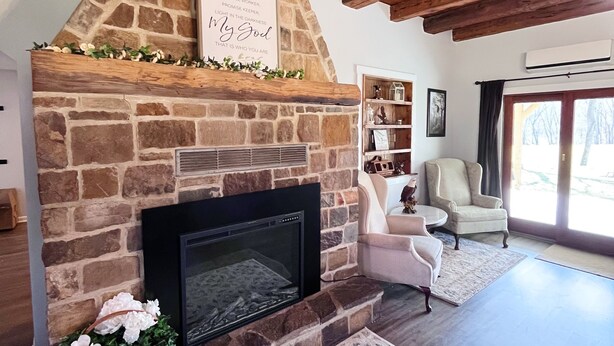 A stone fireplace with a decorative sign, a cozy seating area with a chair and a small table, and a large window providing natural light. - The River House: stone section built in 1790, borders the scenic Juniata  River! (Mifflintown)