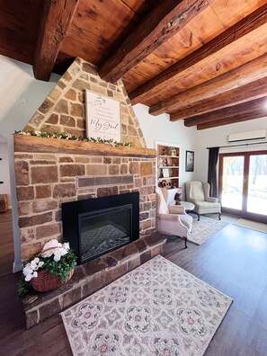 Une cheminée en pierre agrémentée d’une plaque décorative, un coin salon confortable avec un fauteuil et une petite table, et une grande fenêtre laissant entrer la lumière naturelle.
