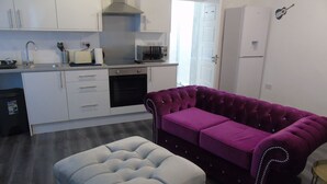 Eine moderne Küche mit weißen Schränken, einem eingebauten Backofen und einer Mikrowelle. Im Wohnbereich befindet sich ein violettes Sofa und ein hellgrauer Hocker.