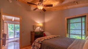 Quarto com teto de madeira, cama com cobertor, mesa de cabeceira com abajur e janela com persianas.