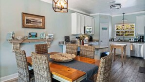 A dining area with a wooden table and wicker chairs, a hanging light fixture, and a kitchen in the background. - Our Place On 30a (Santa Rosa)