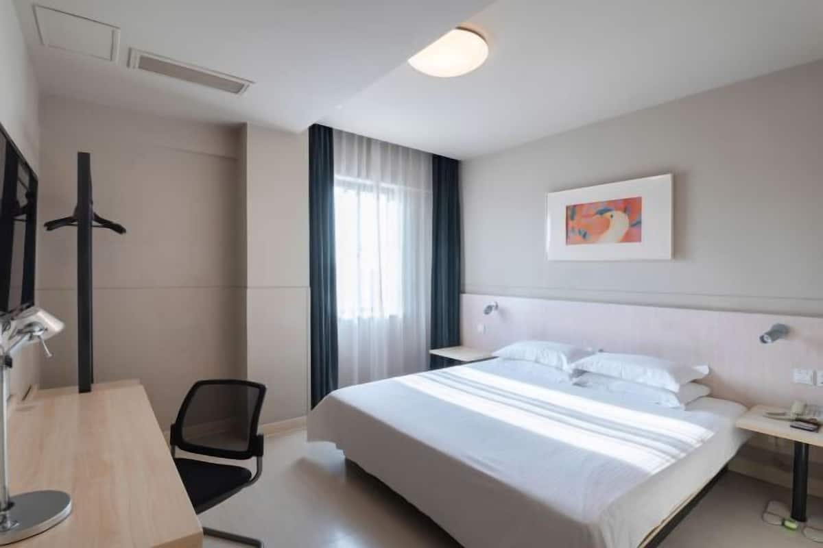 Habitació d'hotel amb llit, escriptori, cadira, televisió, un quadre i finestra amb cortines.