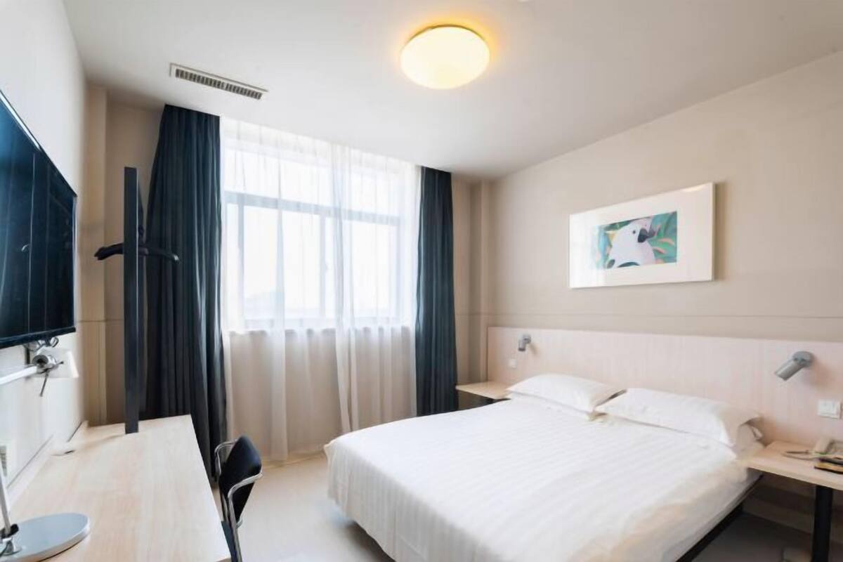 Una habitació d'hotel amb llit, escriptori, cadira, televisió, finestra amb cortines i un quadre a la paret.
