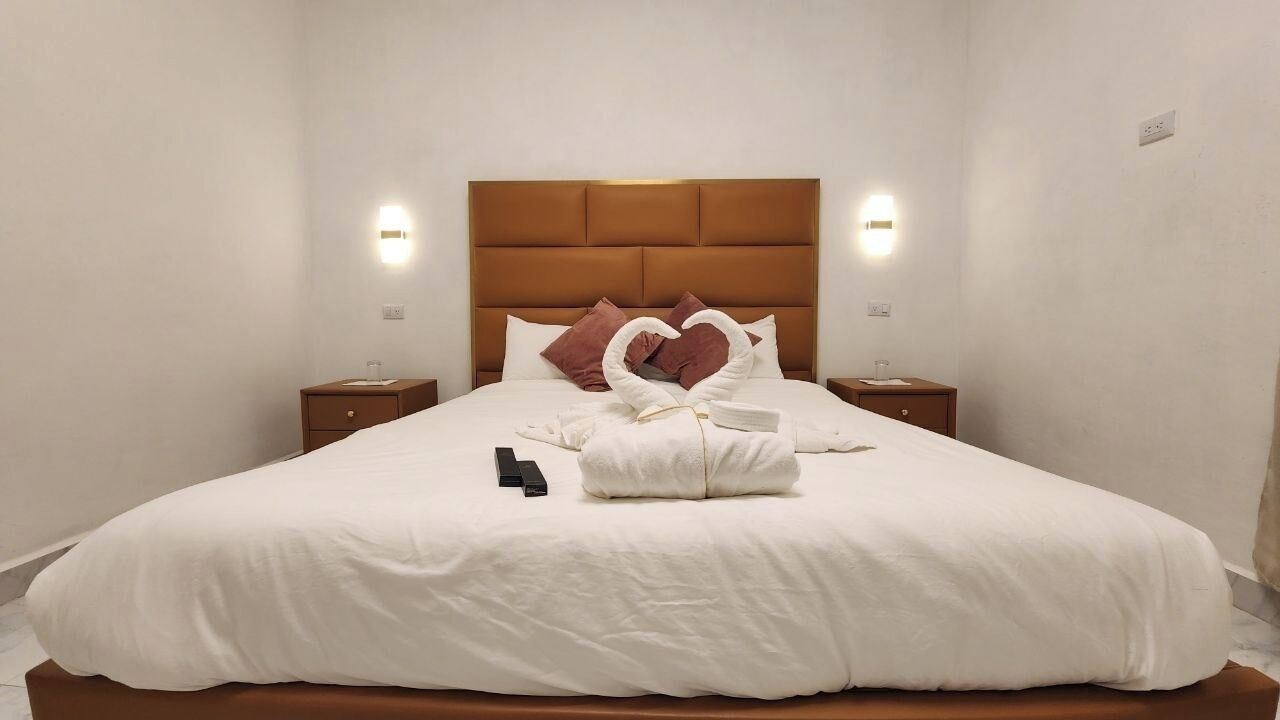A neatly made bed with a brown headboard, white bedding, and decorative swan pillows. There are two bedside tables with lamps and a small nightstand with a lamp.