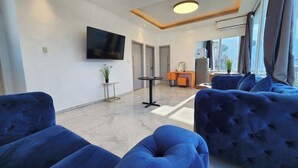 Ein modernes Wohnzimmer mit großem Fernseher, blauen Sofas, einem kleinen runden Tisch und Blick auf die Stadt durch große Fenster.