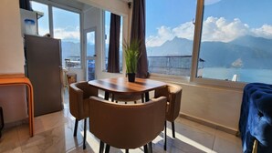 Ein Zimmer mit Esstisch und Stühlen aus Holz, einer kleinen Küchenzeile und einem großen Fenster mit Blick auf Berge und ein Gewässer.