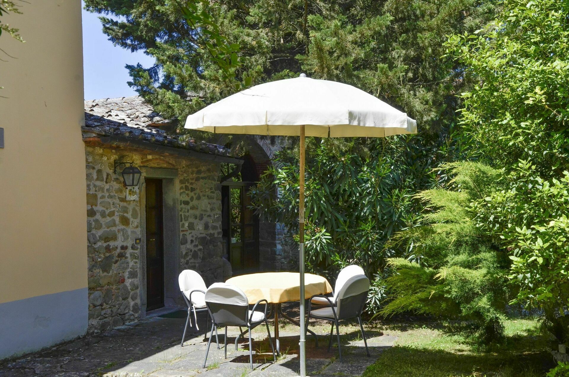 An outdoor patio with a table and chairs under a white umbrella, surrounded by lush greenery and a stone building.