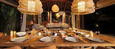 Un área de comedor con una mesa de madera dispuesta para una comida, rodeada de sillas, con un sofá y una mesa de comedor al fondo.