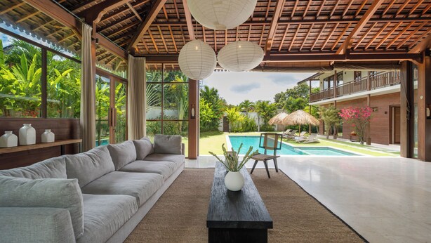 A spacious living area with a large sofa, a coffee table, and a view of a swimming pool and lush garden. - Villa Aloui by Villa Finder (Kerobokan)