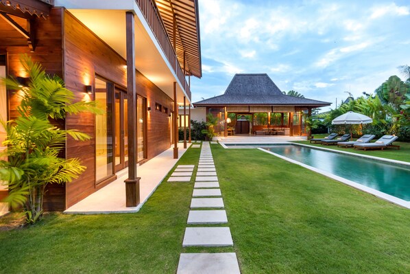 Relax by the pool in a serene outdoor setting, surrounded by lush greenery and a shaded gazebo.