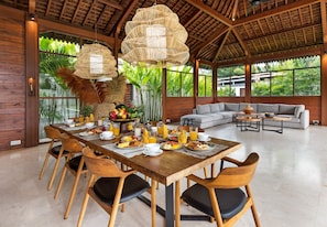 Enjoy breakfast in this spacious dining area with a long table, high ceiling, and large windows offering views of lush greenery.