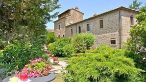 Ein Steinhaus mit einem Turm, umgeben von üppiger Vegetation und einem Garten mit Blumen sowie einem Tisch mit Getränken.