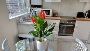 A modern kitchen with white cabinets, a microwave, a toaster, and a sink. There is a glass table with white chairs and a potted plant with red flowers. - Sunny detached bungalow with private courtyard (Bishop's Stortford)