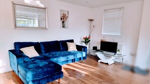 A modern living room with a blue sectional sofa, a flat-screen TV, and a dining area. - Sunny detached bungalow with private courtyard (Bishop's Stortford)