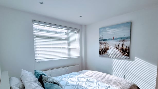A bedroom with a bed, pillows, a radiator, a window with blinds, and a painting on the wall. - Sunny detached bungalow with private courtyard (Bishop's Stortford)