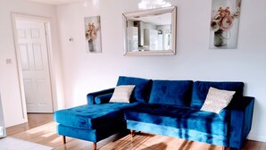 A modern living room with a blue sectional sofa, wooden flooring, a white door, and a large mirror on the wall. - Sunny detached bungalow with private courtyard (Bishop's Stortford)