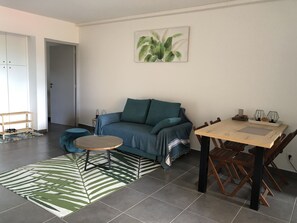 Un salon avec un canapé bleu, une table basse ronde, une table à manger avec des chaises, un mur décoré d’une plante verte et un tapis à motifs.